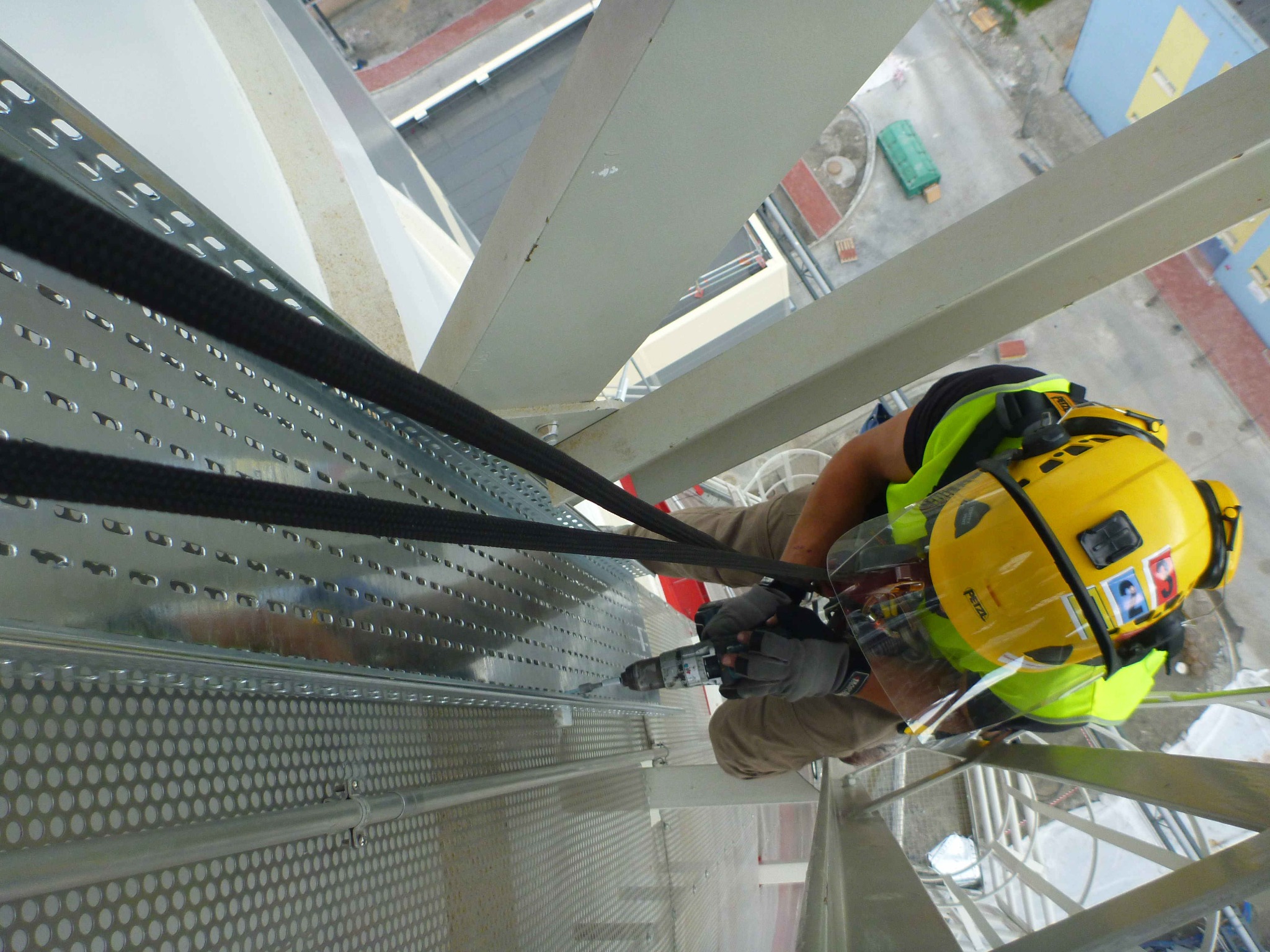 Pracownik w uprzęży i kasku, zabezpieczony linami, wierci w metalowej konstrukcji na wysokości, widok z góry na miasto