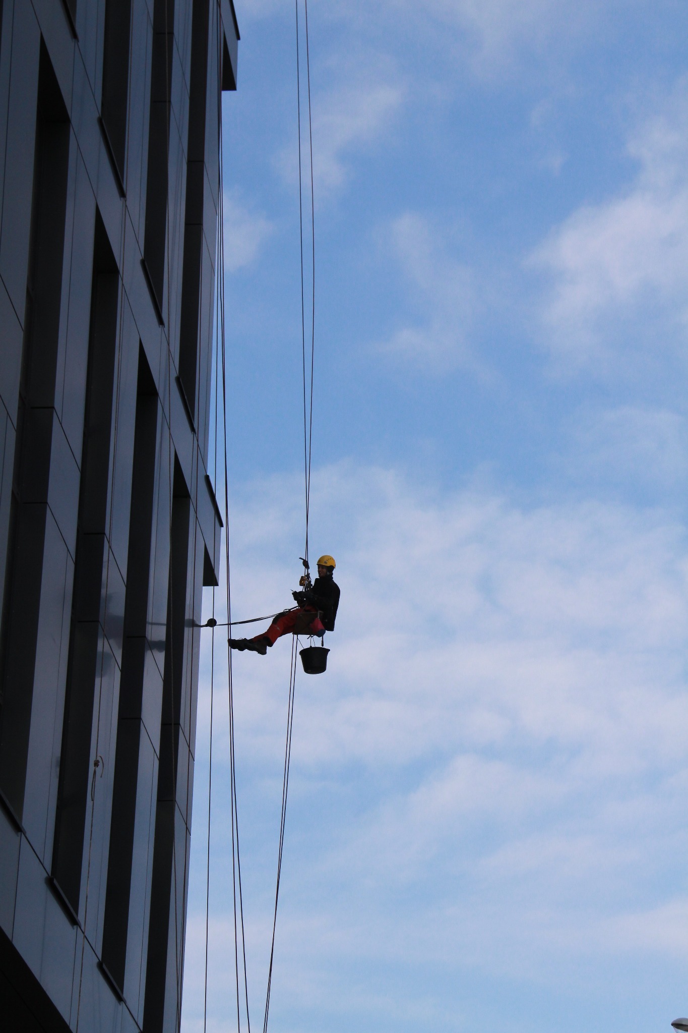 Pracownik na linach, w żółtym kasku, wykonuje prace na wysokości na tle szklanej fasady budynku i błękitnego nieba z chmurami.