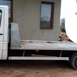 Biała laweta Volkswagen LT 35 z brudnymi kołami stoi na ziemi, w tle budynek z brązowym oknem i reklama usług transportowych z numerem telefonu.