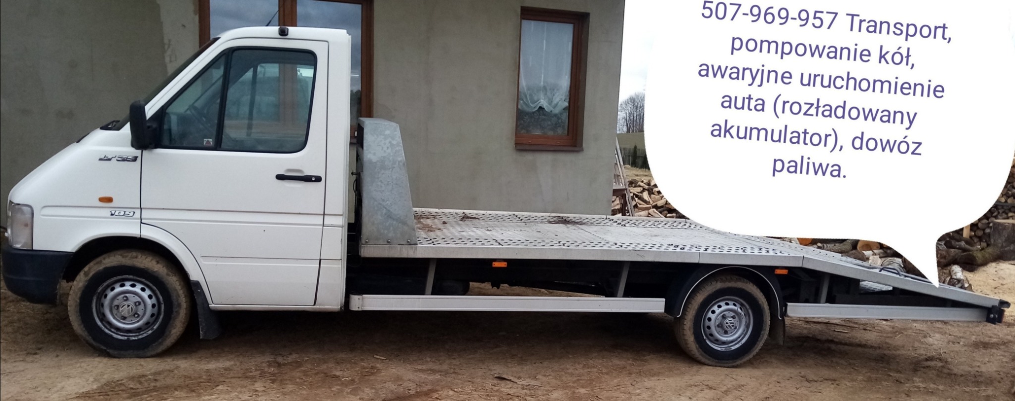 Biała laweta Volkswagen LT 35 z brudnymi kołami stoi na ziemi, w tle budynek z brązowym oknem i reklama usług transportowych z numerem telefonu.