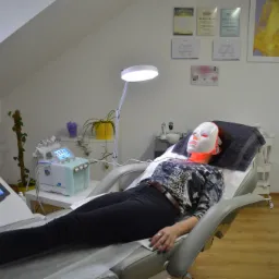 Klientka relaksuje się na fotelu kosmetycznym podczas zabiegu z maską LED, obok widoczny sprzęt do nowoczesnej kosmetyki.