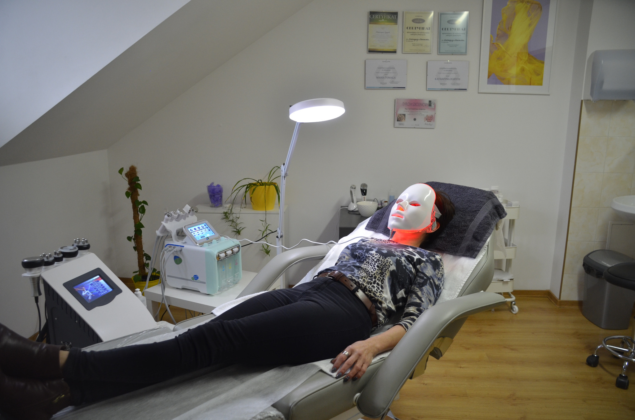 Klientka relaksuje się na fotelu kosmetycznym podczas zabiegu z maską LED, obok widoczny sprzęt do nowoczesnej kosmetyki.