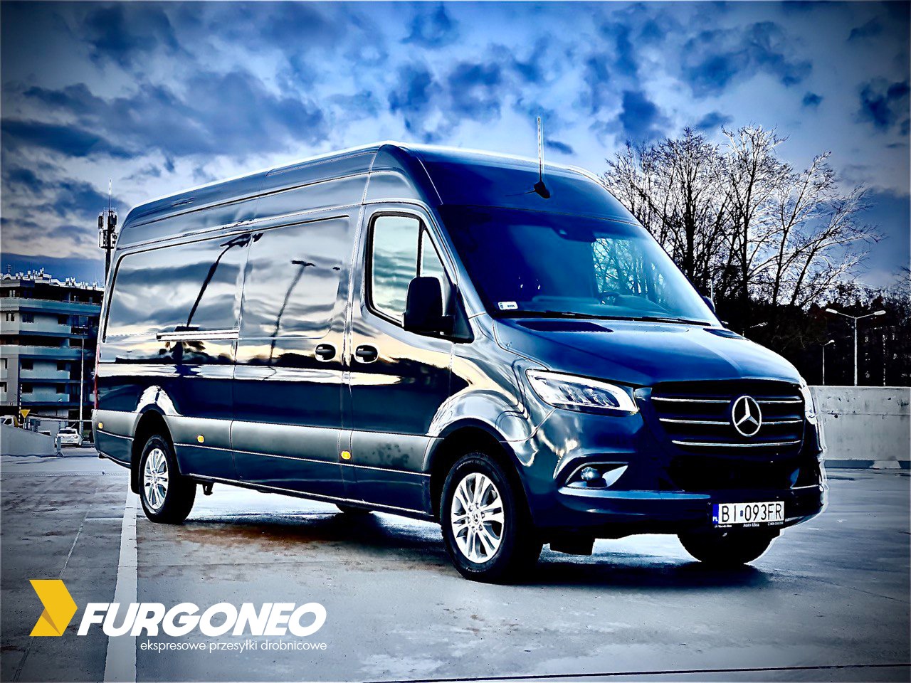 Ciemnogranatowy bus marki Mercedes-Benz Sprinter, zaparkowany na betonowym placu, z widocznym logo firmy przewozowej Furgoneo w lewym dolnym rogu i tablicą rejestracyjną BI 093FR. W tle pochmurne...