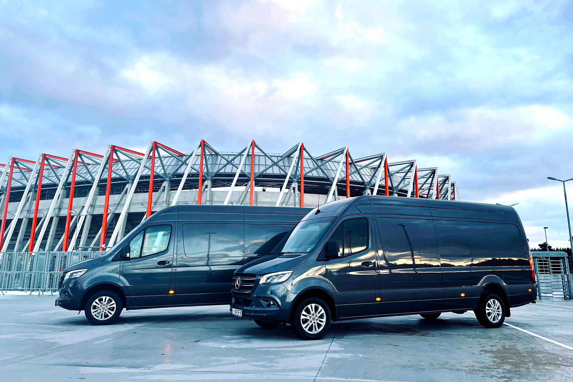 Dwa identyczne, grafitowe busy Mercedes-Benz Sprinter zaparkowane na betonowym placu z widokiem na stadion z czerwonymi elementami konstrukcyjnymi w tle.