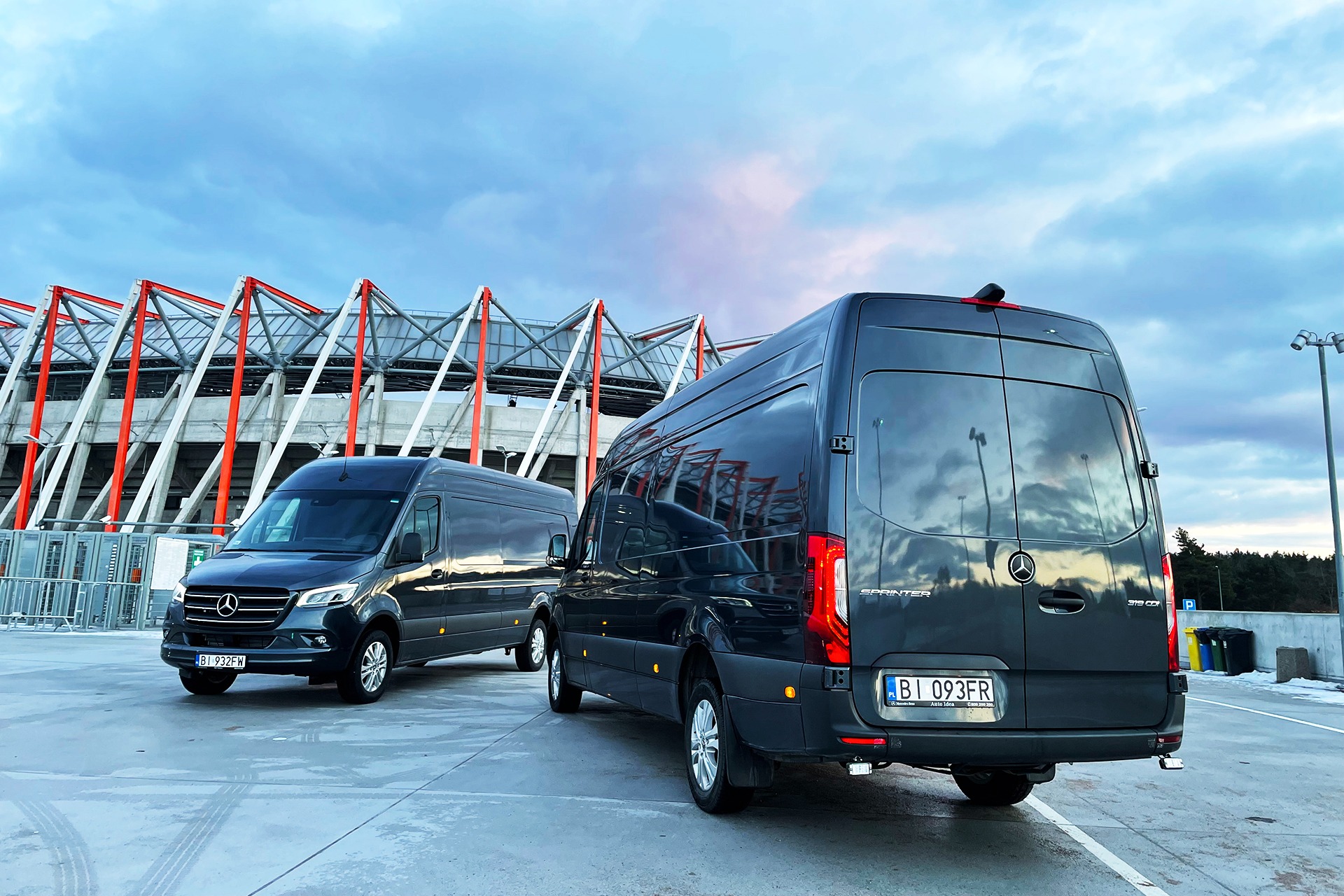 Dwa grafitowe busy Mercedes Sprinter zaparkowane na betonowym placu przed stadionem, widok z tyłu jednego z busów, pochmurne niebo.