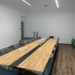 Długi, drewniany stół konferencyjny z czarnym panelem centralnym i szarymi krzesłami w jasnym biurze z białą ceglaną ścianą, nowoczesnym oświetleniem i dekoracyjnymi wazonami.