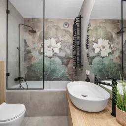 Nowoczesna łazienka z wanną za szklaną ścianką, umywalką nablatową na drewnianym blacie i tapetą w kwiaty lotosu, oświetlona ciepłym światłem.
