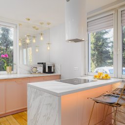 Dream Home Estate Sp. z o.o. - Jasna kuchnia z różowymi szafkami, marmurowym blatem, złotą armaturą i krzesłami, oświetlona wiszącymi lampami i światłem dziennym z okien z widokiem na zieleń.