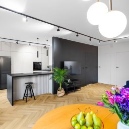 Dream Home Estate Sp. z o.o. - Nowoczesne wnętrze mieszkania z parkietem w jodełkę, wyspą kuchenną z hokerem, czarną ścianą telewizyjną i bukietem kwiatów na pierwszym planie.