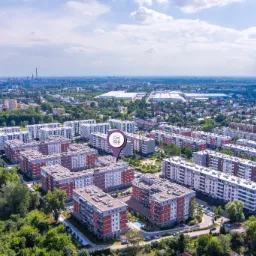 Panorama osiedla mieszkaniowego z lotu ptaka, widoczny kompleks budynków z czerwono-szarymi elewacjami, otoczony zielenią drzew, w tle panorama miasta z drapaczami chmur, na pierwszym planie...