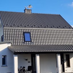 Dream Home Estate Sp. z o.o. - Dom z ciemnoszarą dachówką ceramiczną i ozdobną wiatrowskaz na kominie. Widoczna antena satelitarna na białej ścianie budynku.