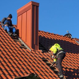 Dream Home Estate Sp. z o.o. - Dwóch dekarzy na dachu z czerwonej dachówki. Jeden siedzi obok komina, drugi naprawia dach, używając wkrętarki. Błękitne niebo w tle.