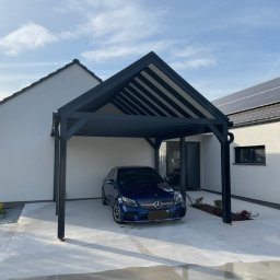 Dream Home Estate Sp. z o.o. - Nowoczesna, grafitowa wiata garażowa z zadaszeniem lamelowym, przylegająca do białego domu z panelami słonecznymi na dachu. Pod wiatą stoi ciemnoniebieski samochód.