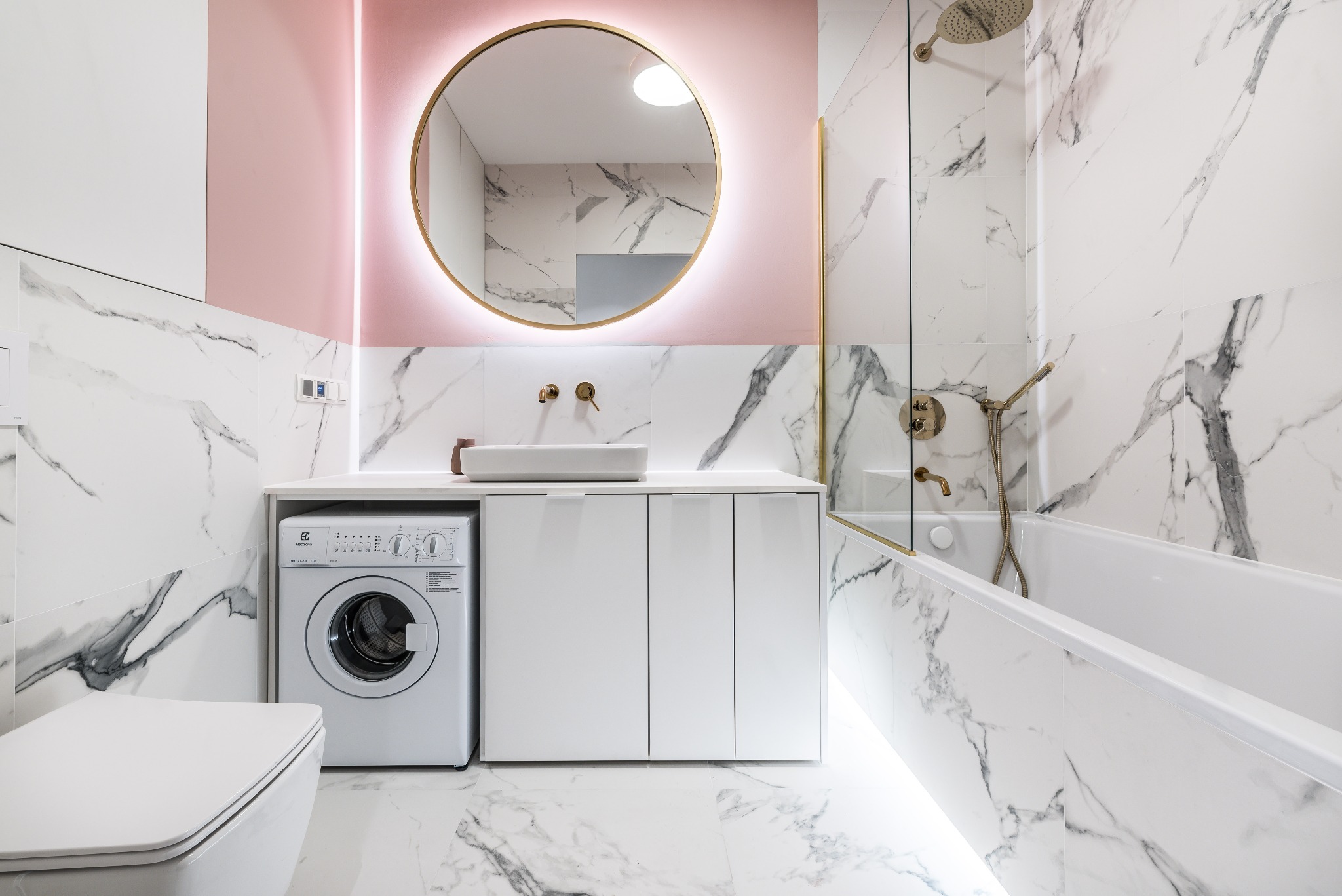 Elegancka łazienka z marmurowymi płytkami, okrągłym lustrem z podświetleniem LED, białą umywalką nablatową, pralką Electrolux i kabiną prysznicową ze złotymi akcentami.