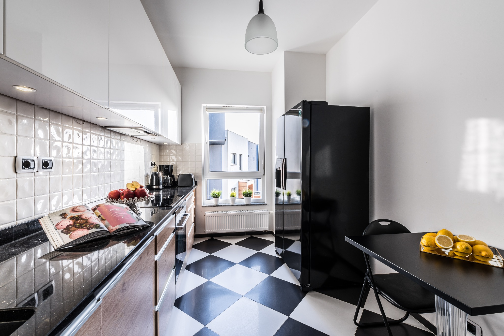 Nowoczesna kuchnia z czarno-białą podłogą w szachownicę, czarny blat, białe szafki, czarna lodówka, stół z cytrynami i książka kucharska na blacie.