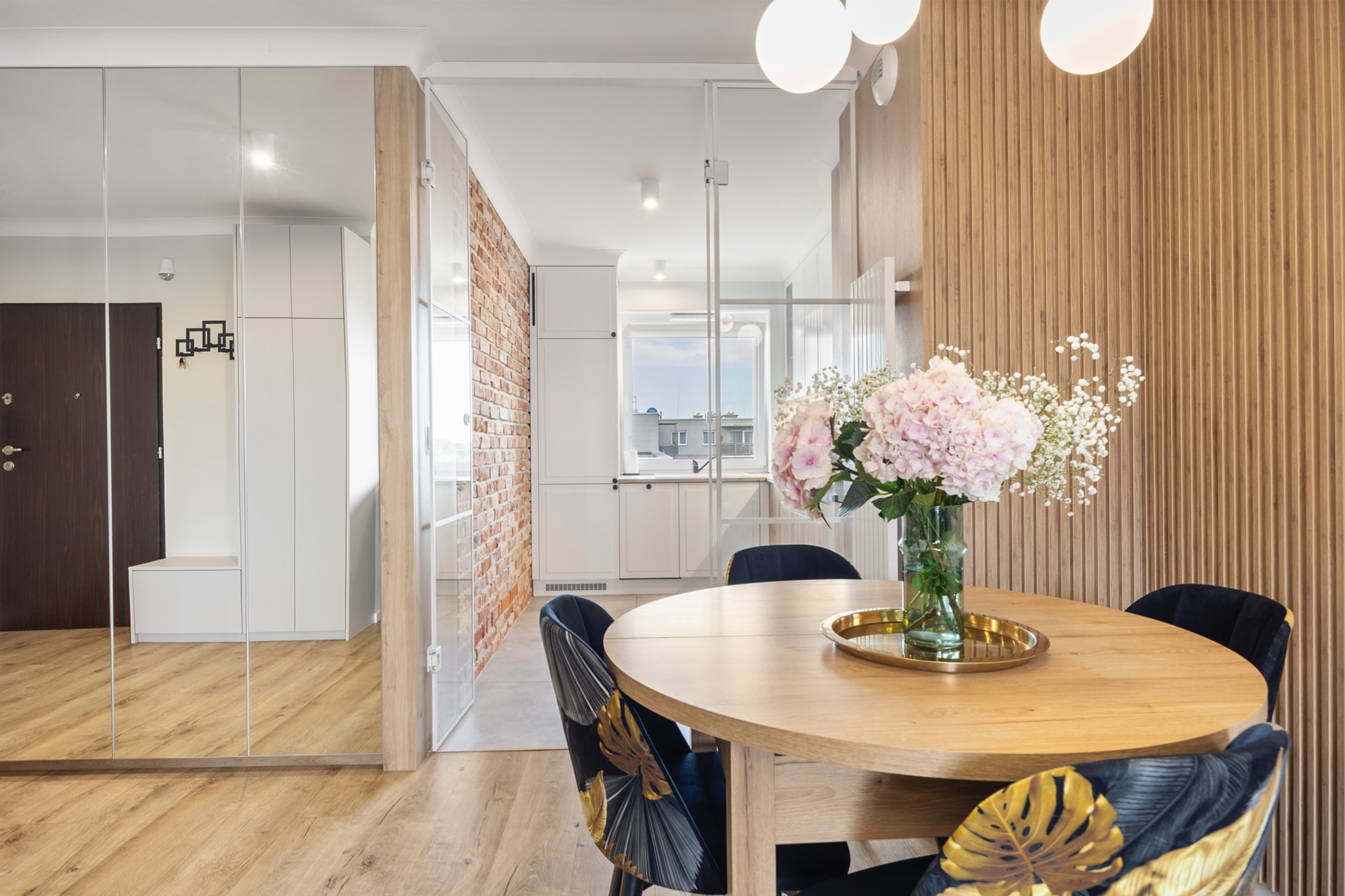 Jasne wnętrze mieszkania z okrągłym drewnianym stołem, bukietem różowych hortensji i dekoracyjnymi krzesłami na tle lustrzanej ściany i ceglanej ściany za szklanymi drzwiami.