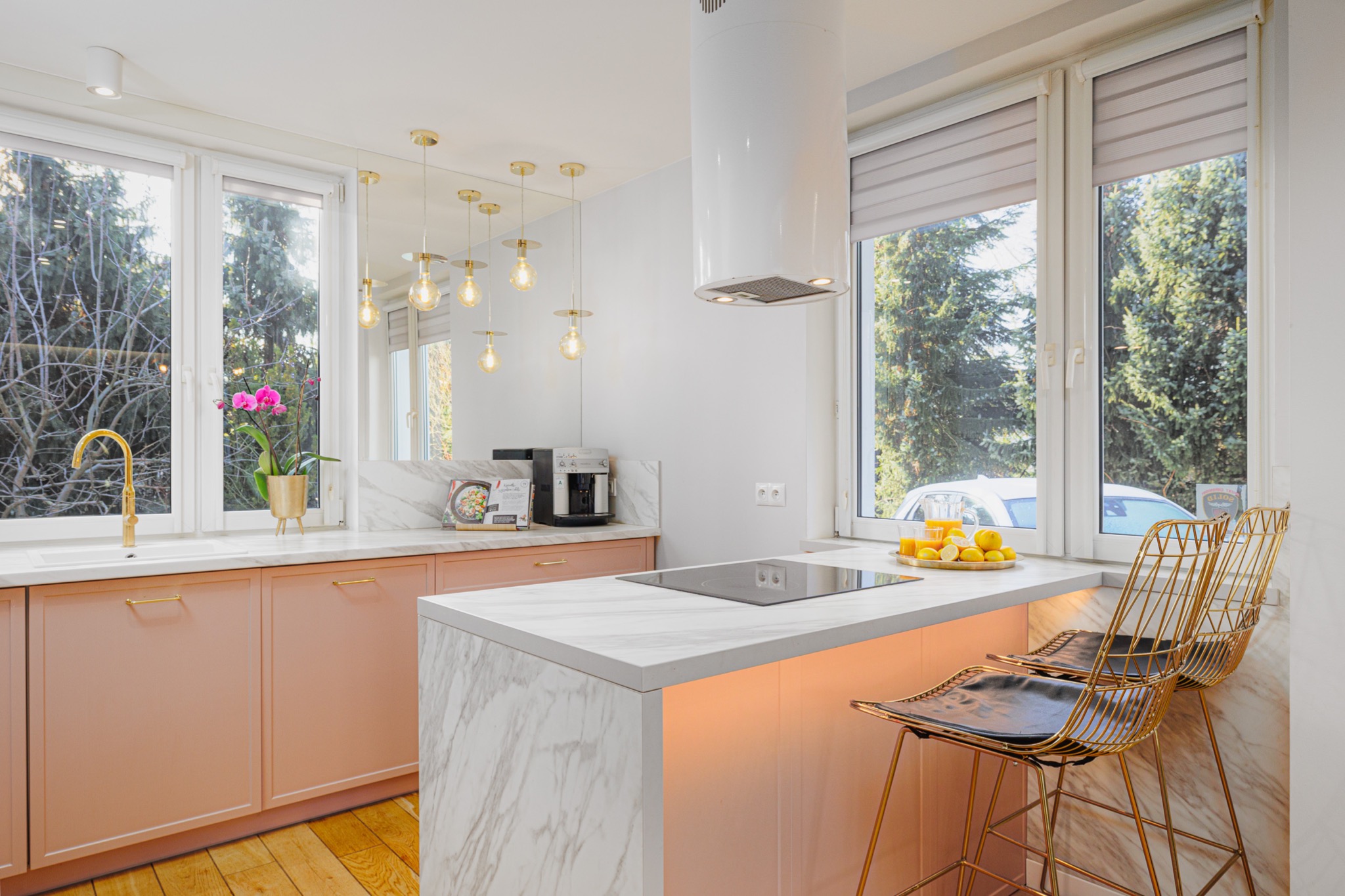 Jasna kuchnia z różowymi szafkami, marmurowym blatem, złotą armaturą i krzesłami, oświetlona wiszącymi lampami i światłem dziennym z okien z widokiem na zieleń.