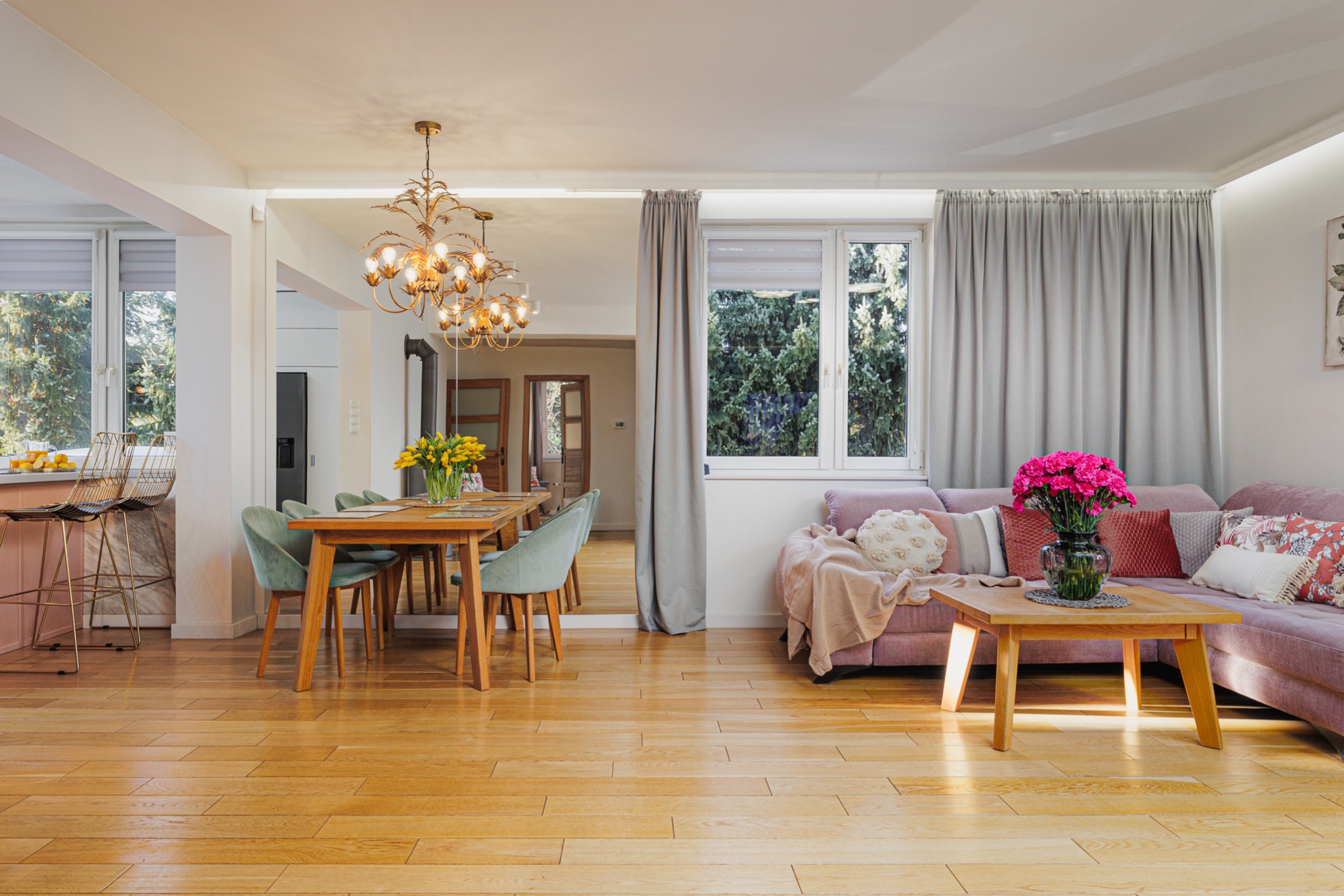 Jasne wnętrze salonu z drewnianą podłogą, stołem jadalnym z zielonymi krzesłami, sofą w odcieniach fioletu z poduszkami i bukietem różowych kwiatów na niskim stoliku kawowym; w tle widoczna kuchnia...