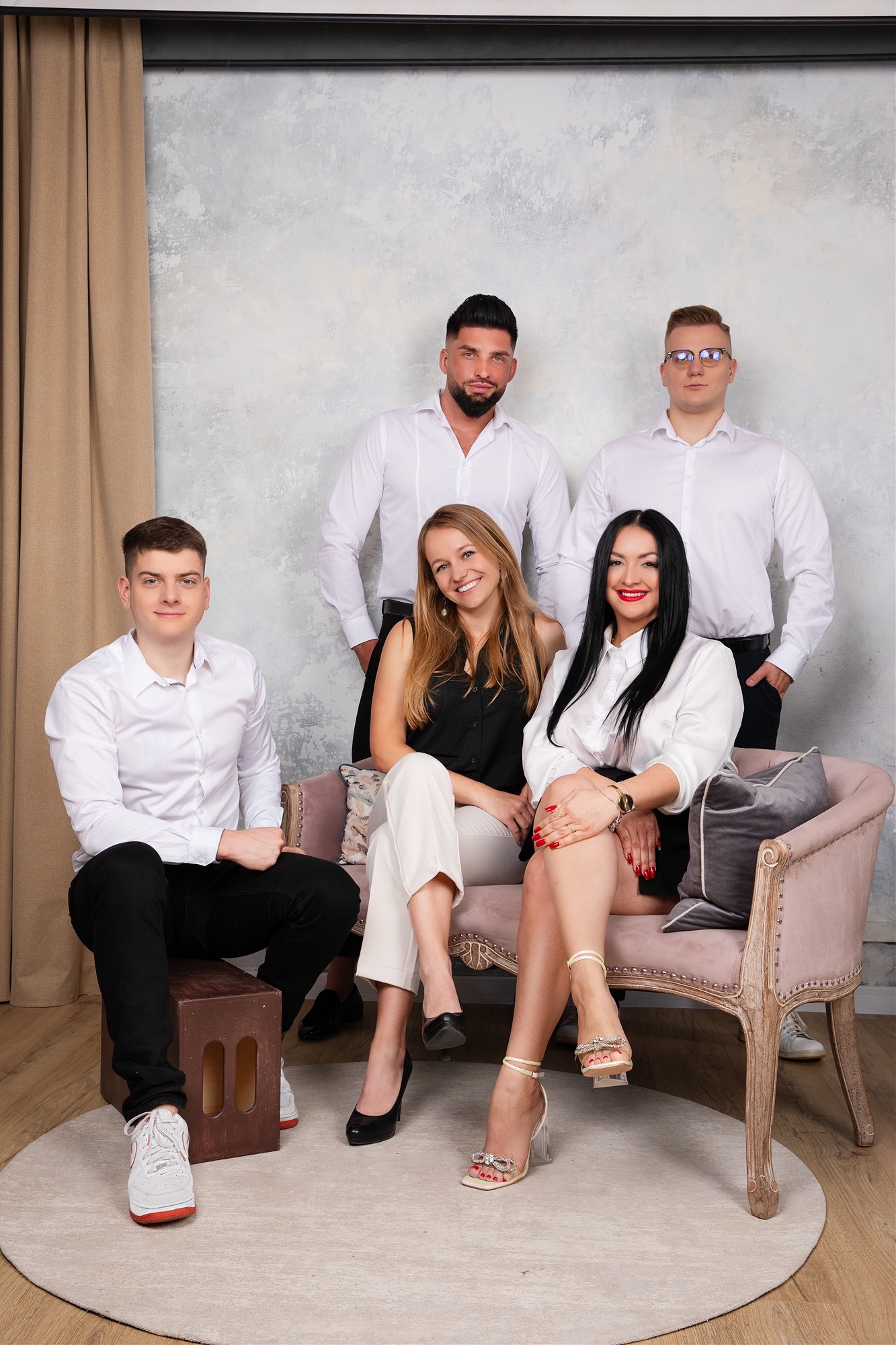 Zespół pracowników agencji nieruchomości pozuje w studio: dwie kobiety siedzą na różowej kanapie, trzej mężczyźni stoją za nimi, wszyscy ubrani w białe koszule. Jeden mężczyzna siedzi na drewnianym...