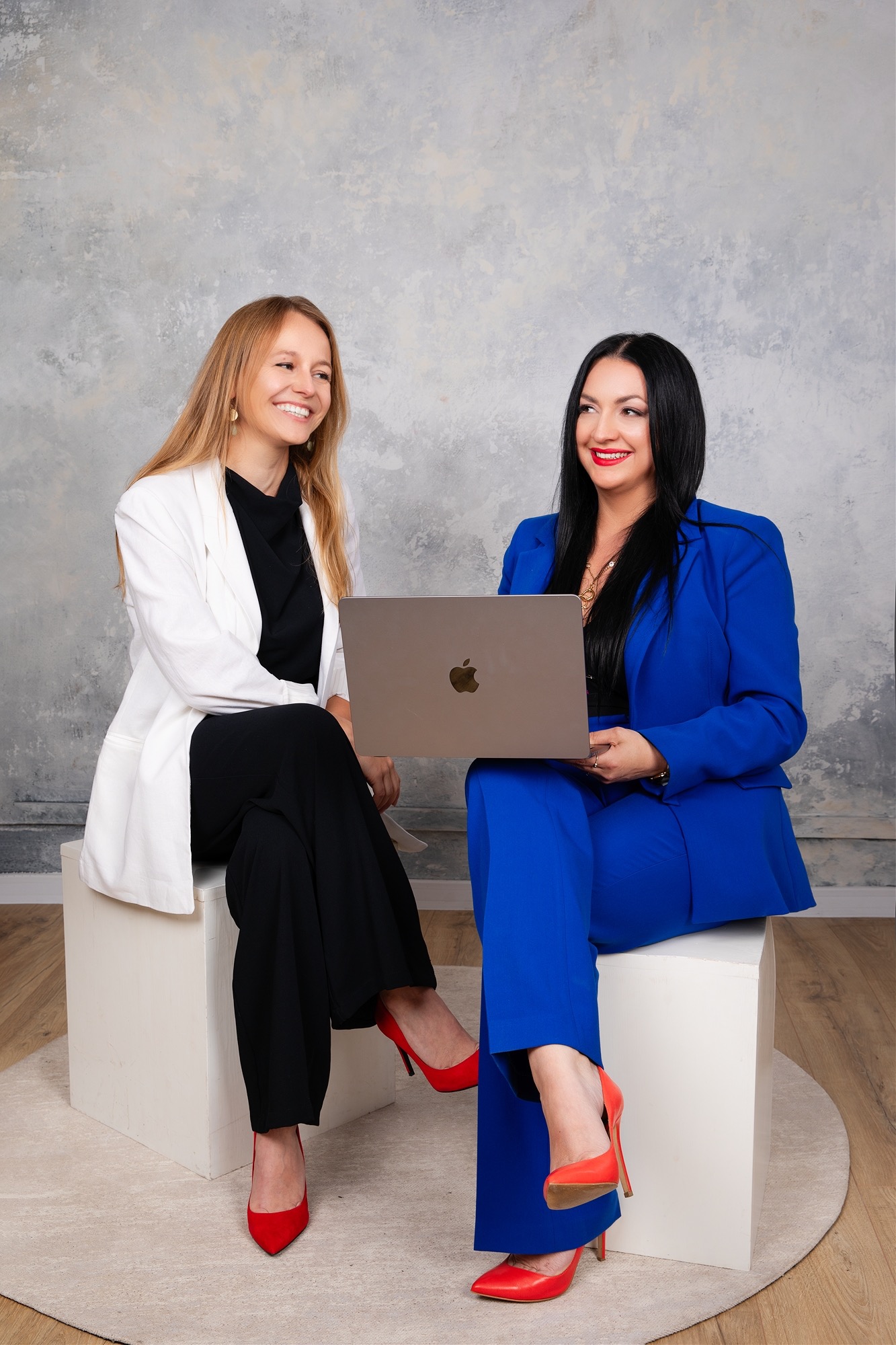 Dwie uśmiechnięte kobiety w eleganckich strojach (biała marynarka i czarne spodnie oraz niebieski garnitur) siedzą na białych kostkach i rozmawiają, jedna trzyma laptopa, obie mają czerwone szpilki...