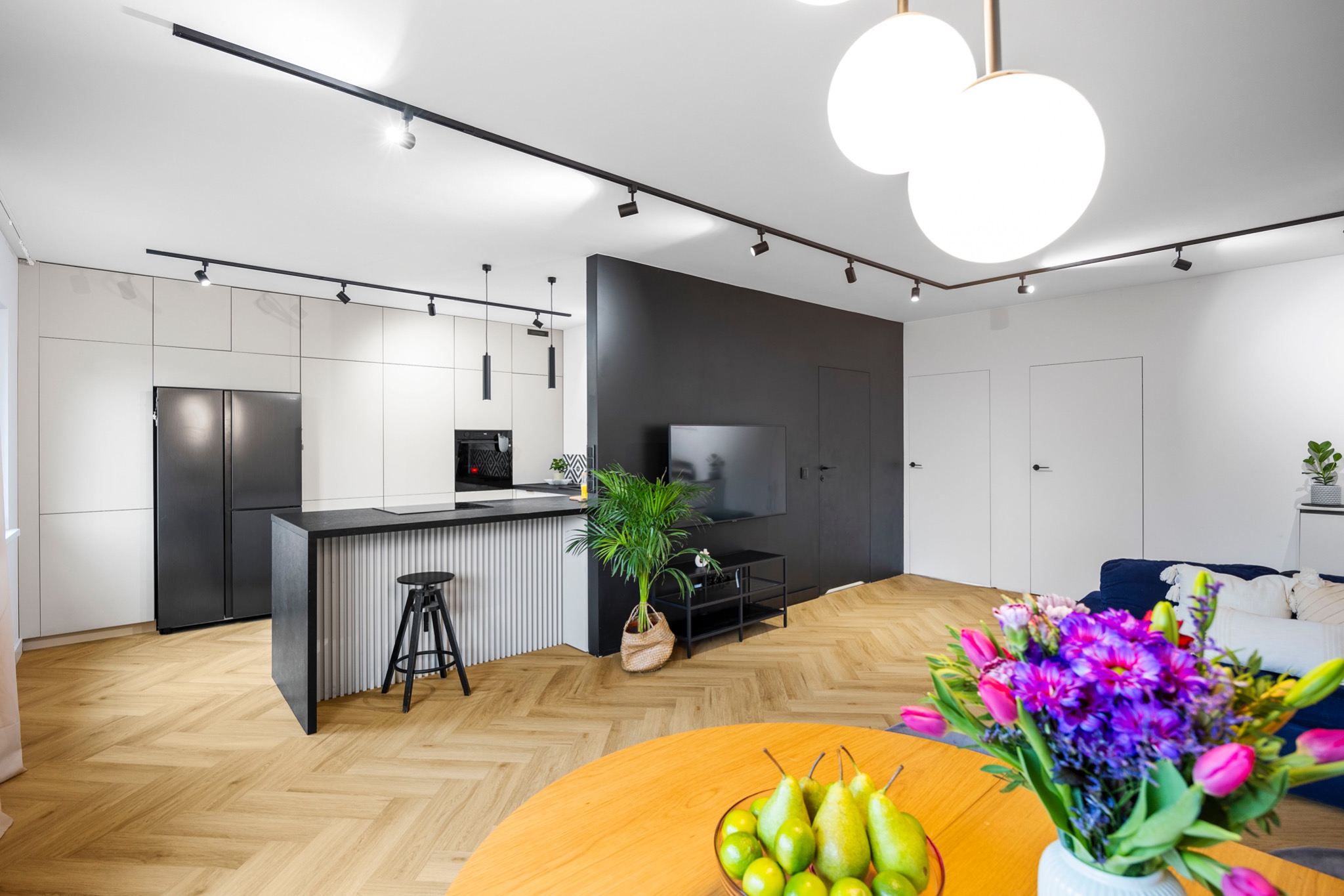 Nowoczesne wnętrze mieszkania z parkietem w jodełkę, wyspą kuchenną z hokerem, czarną ścianą telewizyjną i bukietem kwiatów na pierwszym planie.