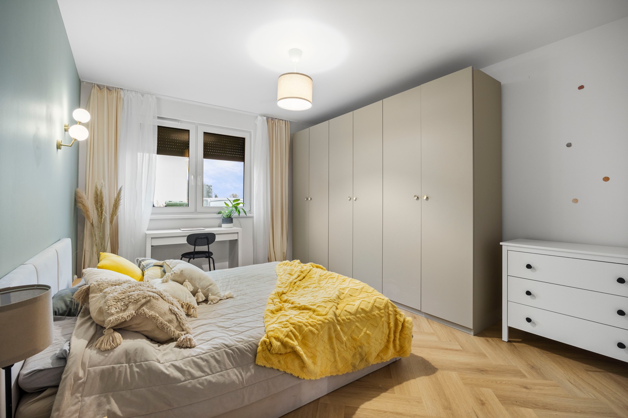 Jasna sypialnia z parkietem w jodełkę, beżową szafą w zabudowie, biurkiem przy oknie i łóżkiem z dekoracyjnymi poduszkami i żółtym pledem.