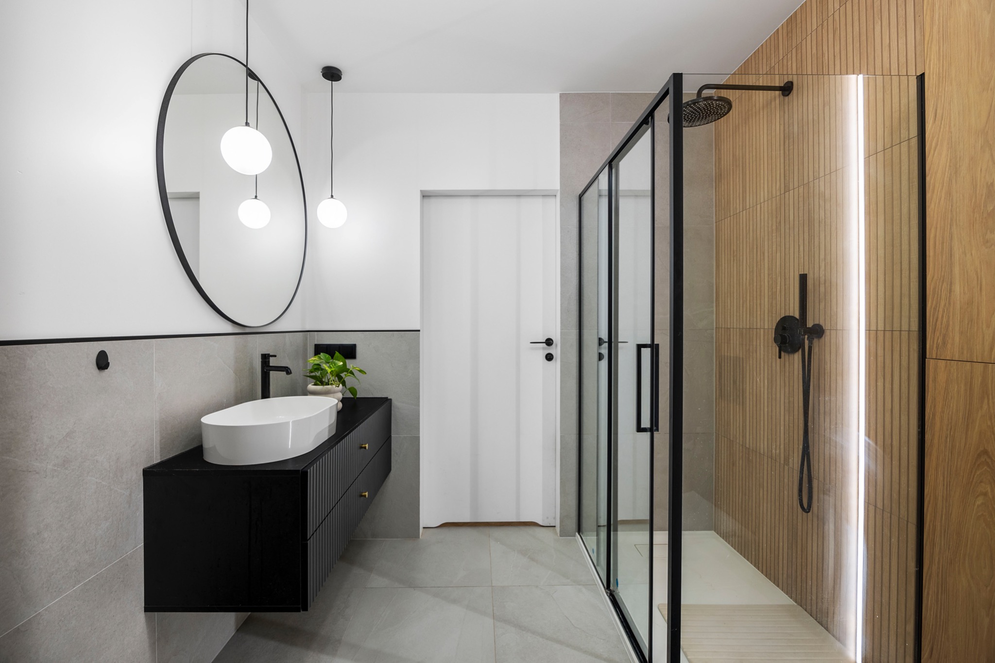 Nowoczesna łazienka z kabiną prysznicową typu walk-in, owalną umywalką na czarnej szafce z frezowanymi frontami, okrągłym lustrem i drewnianą ścianą w kabinie prysznicowej z oświetleniem LED.
