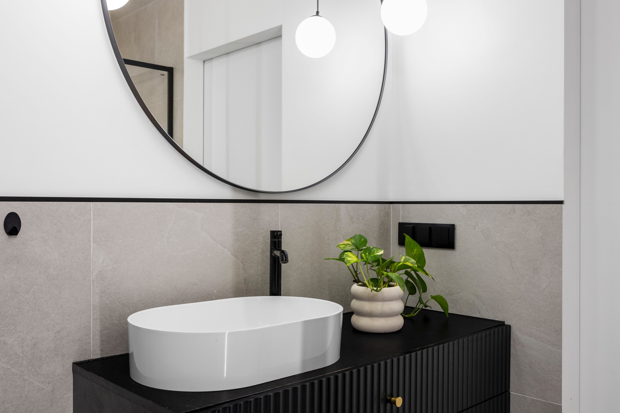 Nowoczesna łazienka z owalną białą umywalką nablatową, czarną armaturą i szafką z frezowanymi frontami, uzupełniona okrągłym lustrem i rośliną doniczkową w ceramicznej osłonce.