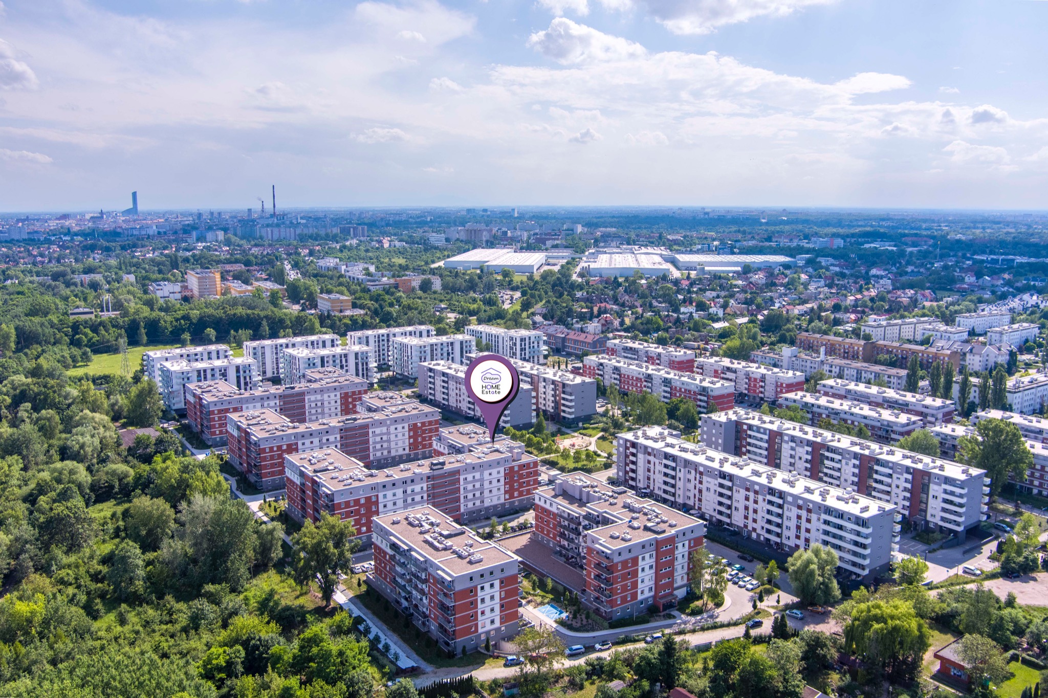 Panorama osiedla mieszkaniowego z lotu ptaka, widoczny kompleks budynków z czerwono-szarymi elewacjami, otoczony zielenią drzew, w tle panorama miasta z drapaczami chmur, na pierwszym planie...