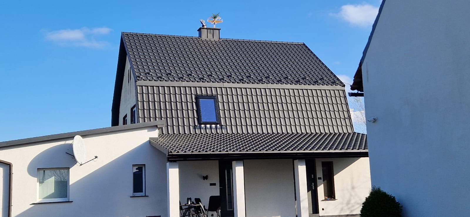 Dom z ciemnoszarą dachówką ceramiczną i ozdobną wiatrowskaz na kominie. Widoczna antena satelitarna na białej ścianie budynku.
