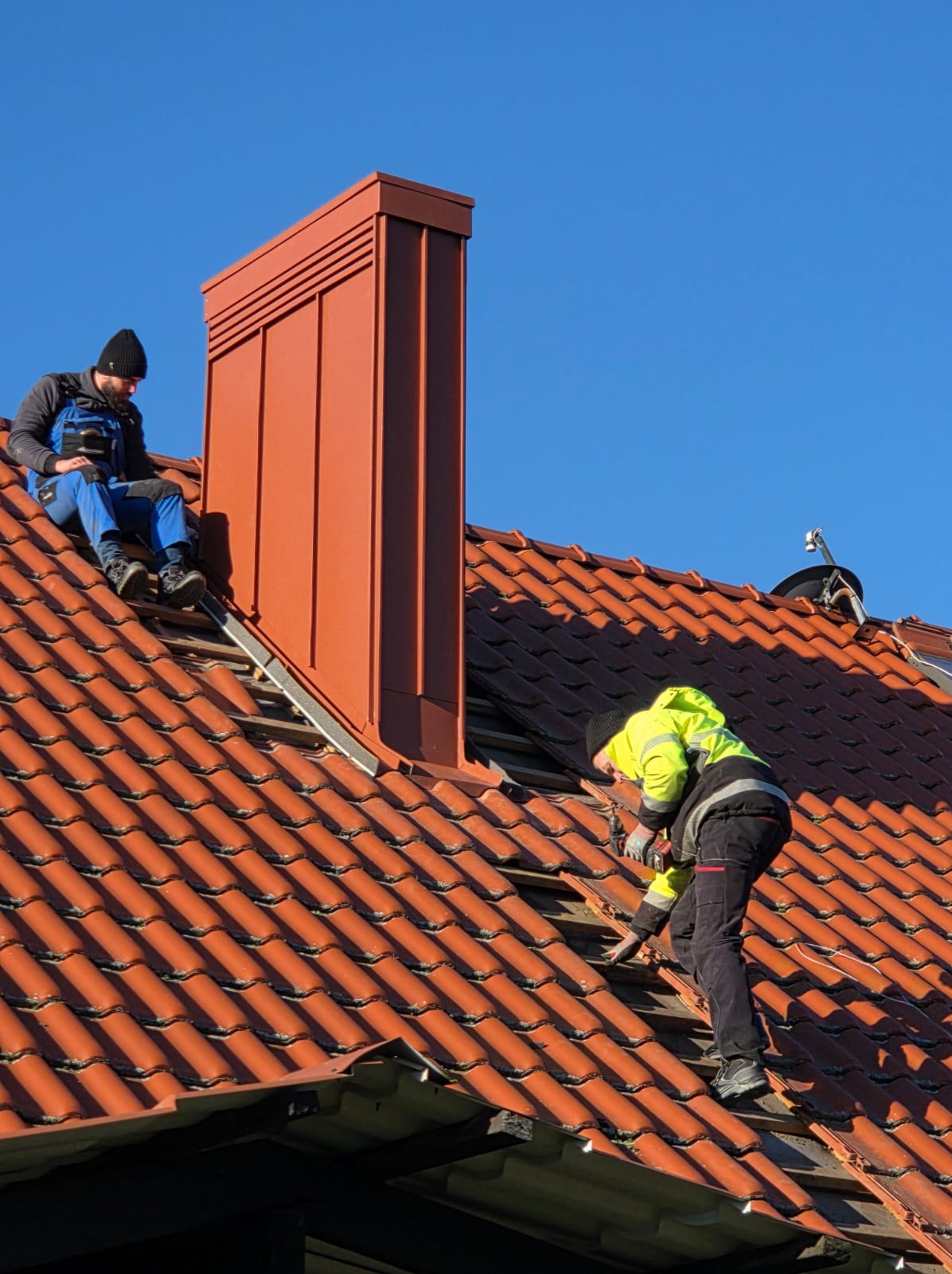 Dwóch dekarzy na dachu z czerwonej dachówki. Jeden siedzi obok komina, drugi naprawia dach, używając wkrętarki. Błękitne niebo w tle.