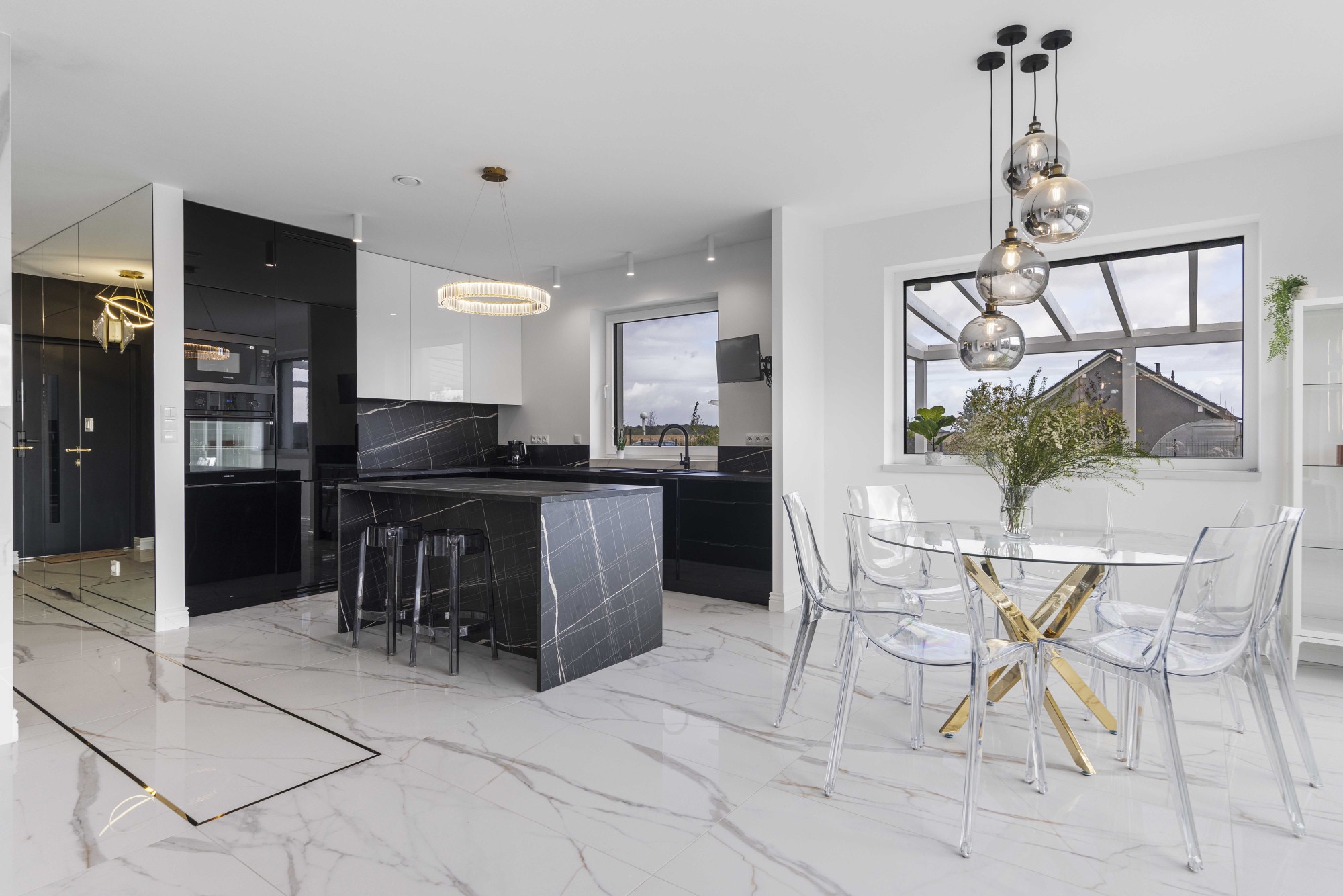 Nowoczesna kuchnia z wyspą o marmurowym wykończeniu, szklanym stołem i przezroczystymi krzesłami. Białe ściany, marmurowa podłoga z czarnymi akcentami.