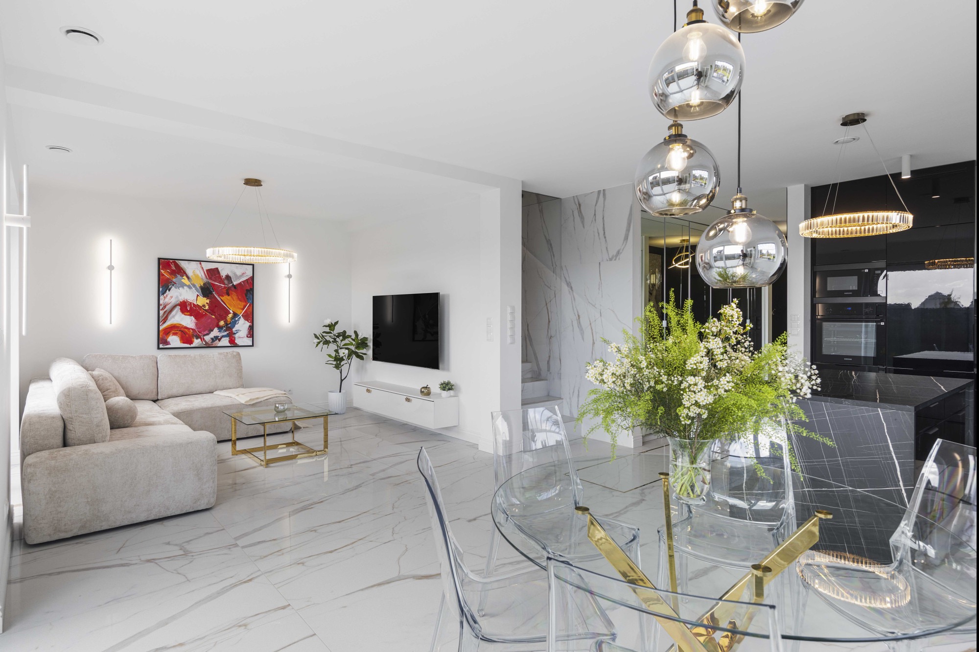 Nowoczesne wnętrze: salon z narożnikiem, szklanym stolikiem i marmurową podłogą, przechodzący w jadalnię ze szklanym stołem i kuchnią z czarnym blatem.