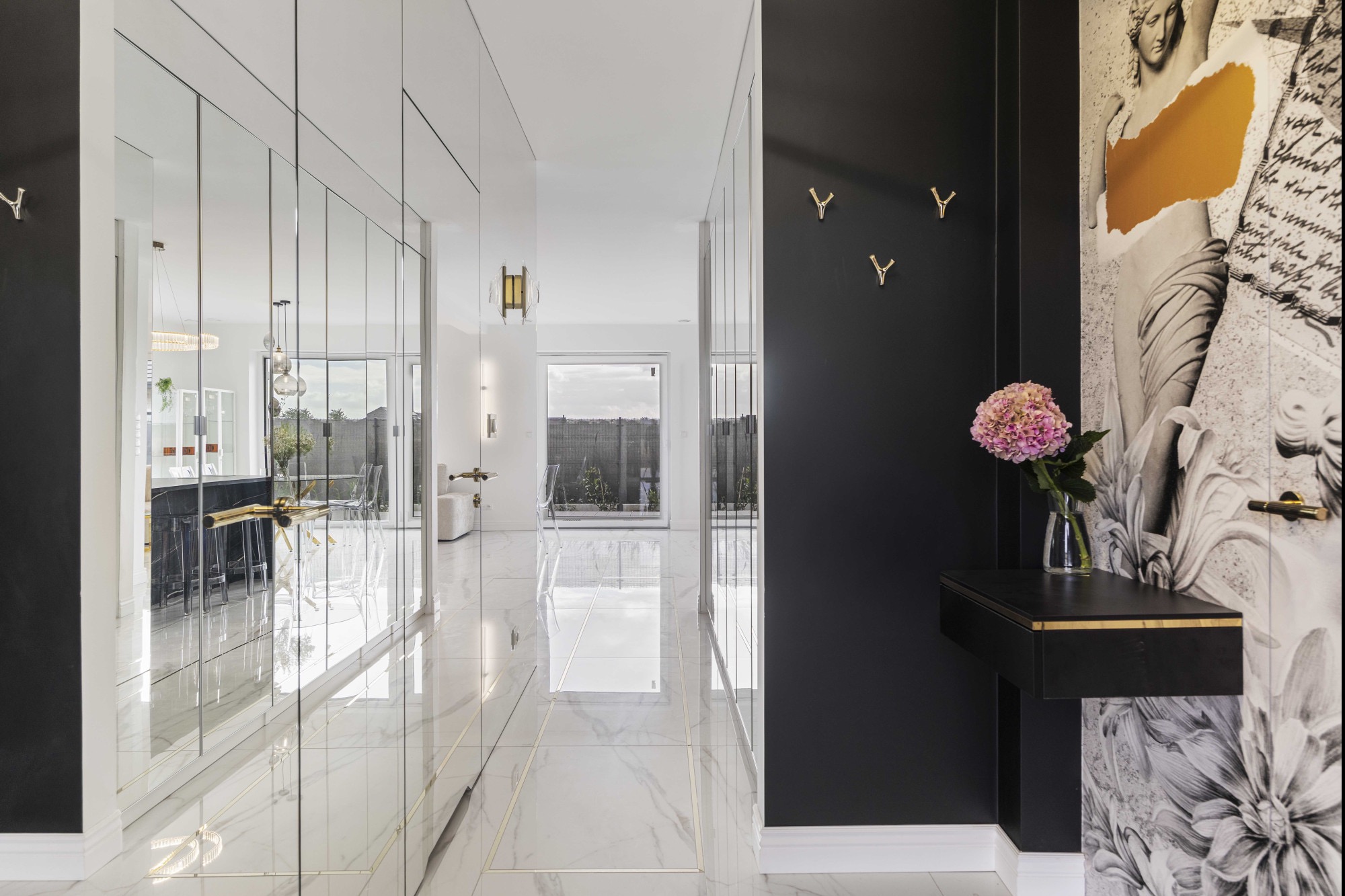 Nowoczesny korytarz z lustrzanymi szafami, marmurową podłogą i czarną ścianą z dekoracyjnym muralem oraz złotymi akcentami. Widok na salon i ogród.