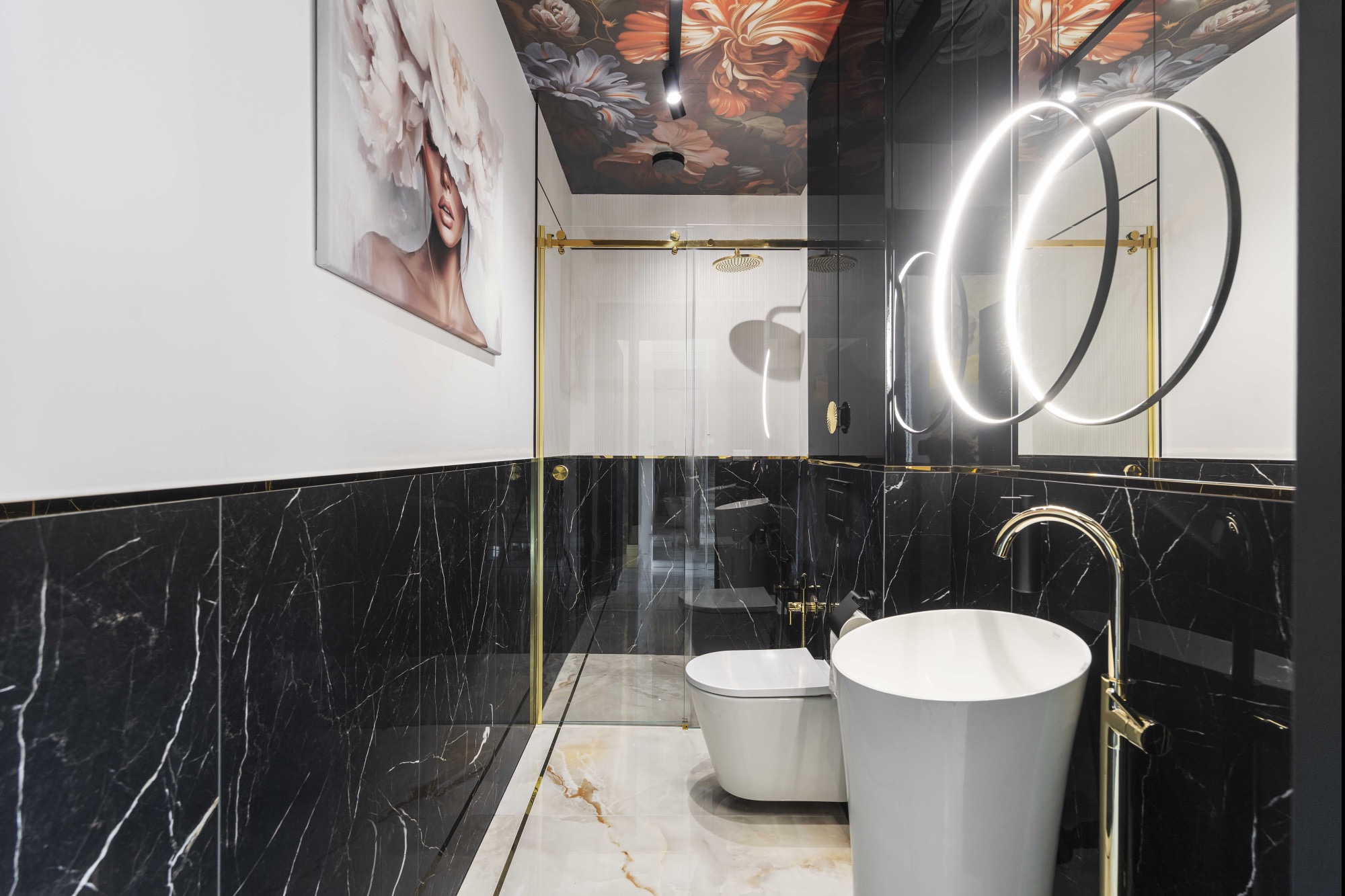 Elegancka łazienka z czarnymi marmurowymi płytkami, złotymi akcentami i nowoczesnym oświetleniem. Na ścianie obraz z motywem kwiatowym, a na suficie tapeta w kwiaty.