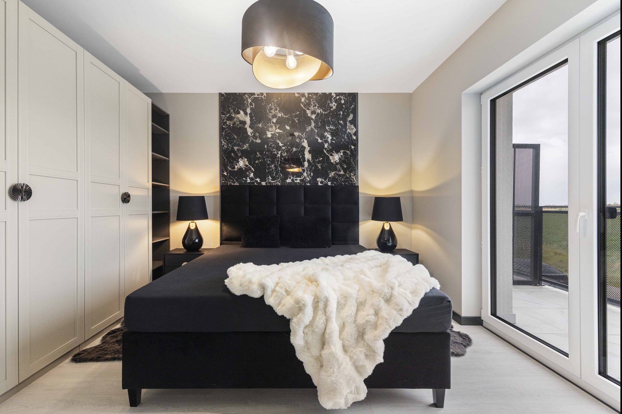 Elegancka sypialnia z czarnym łóżkiem, marmurową ścianą i wyjściem na balkon. Biała szafa w zabudowie i designerska lampa sufitowa dopełniają wystrój.