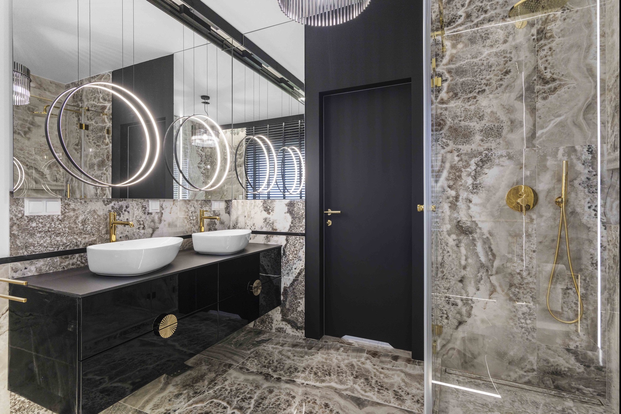Elegancka łazienka z marmurowymi płytkami, czarną szafką pod umywalki i złotymi akcentami, oświetlona okrągłymi lampami. Widoczna kabina prysznicowa.