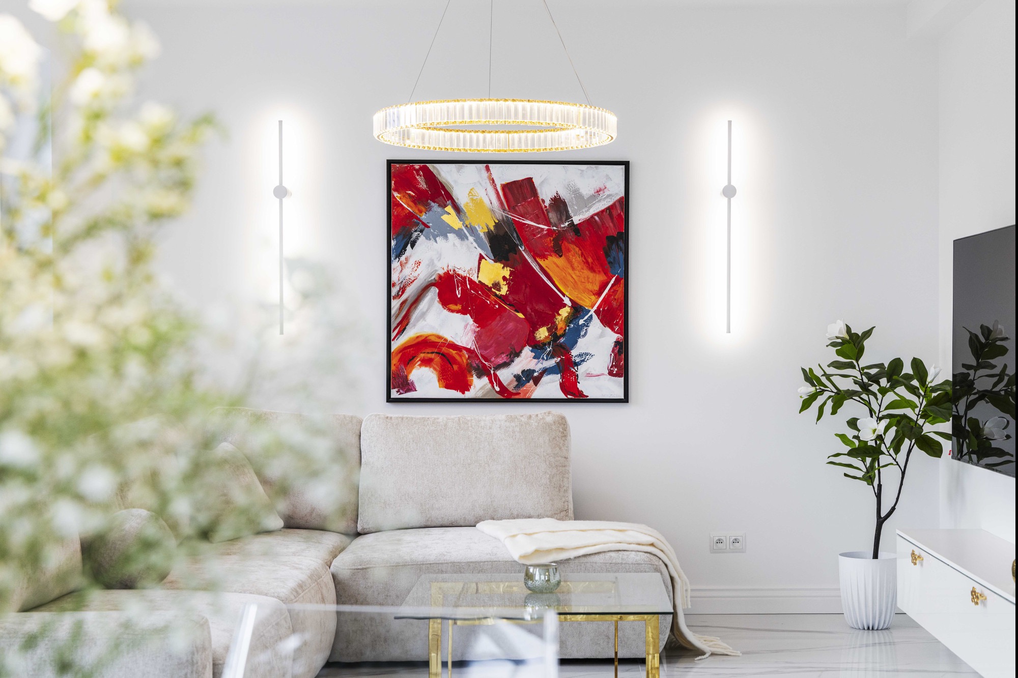 Nowoczesny salon z narożnikiem, abstrakcyjnym obrazem w czarnej ramie, okrągłą lampą i minimalistycznymi kinkietami. Jasne wnętrze z marmurową podłogą i roślinami.