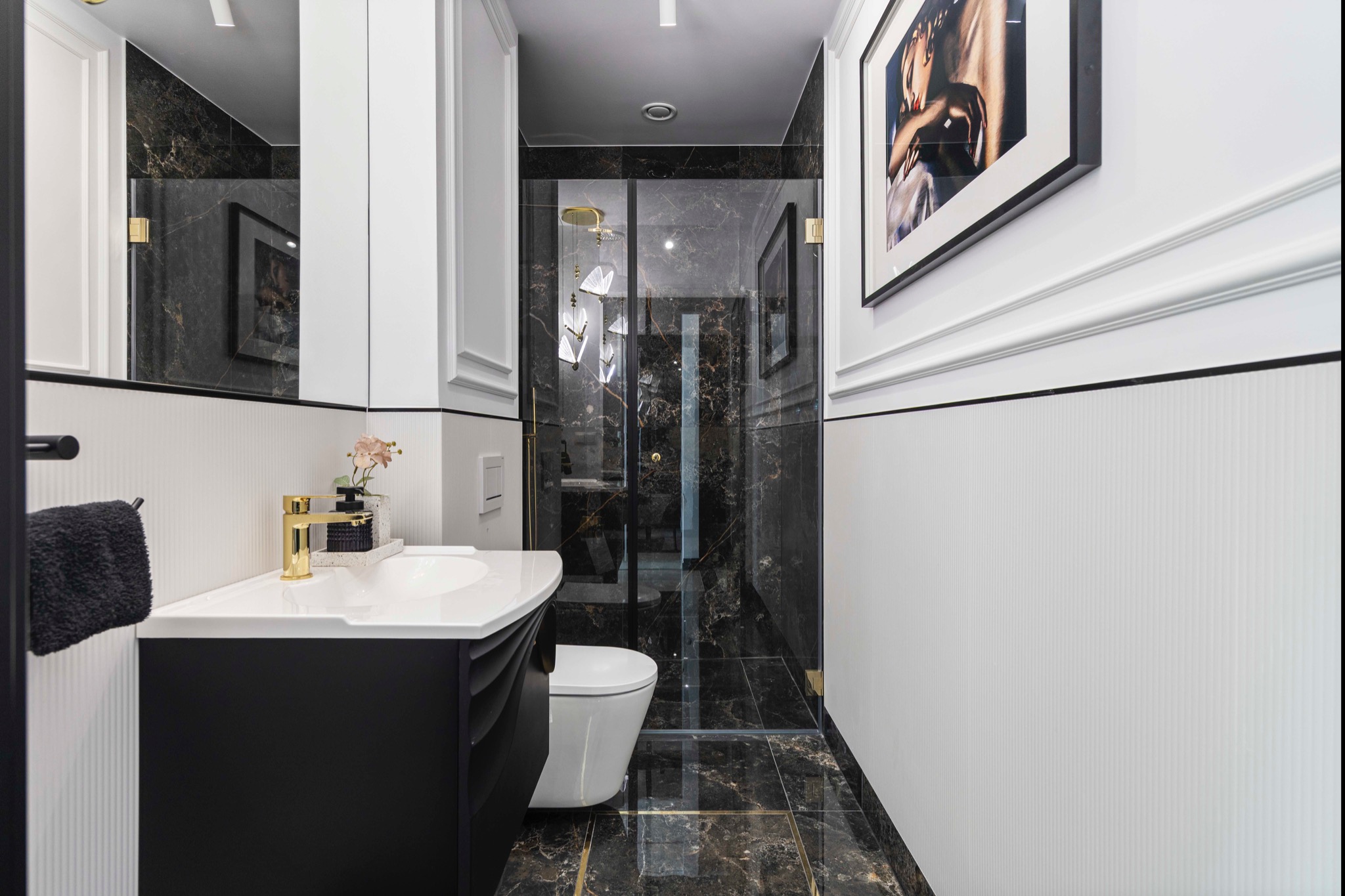 Elegancka łazienka w czerni i bieli z marmurowymi akcentami, złotą armaturą i dekoracyjnym oświetleniem. Widoczna umywalka, toaleta i kabina prysznicowa.