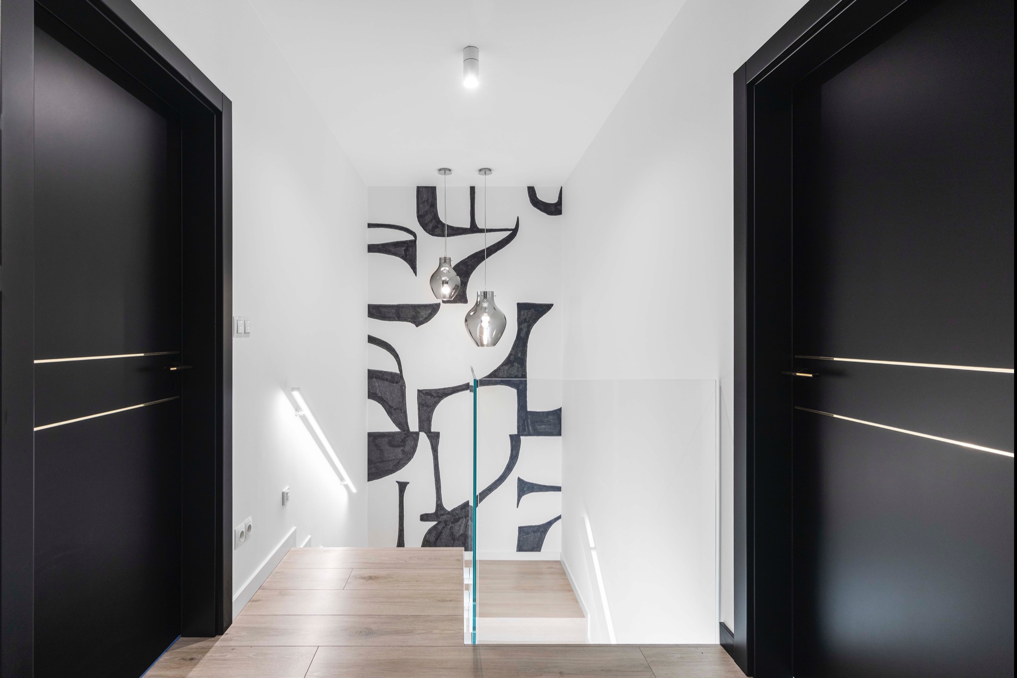 Nowoczesne wnętrze z czarnymi drzwiami i abstrakcyjną grafiką na ścianie we Wrocławiu. Szklana balustrada i drewniane schody dodają elegancji.