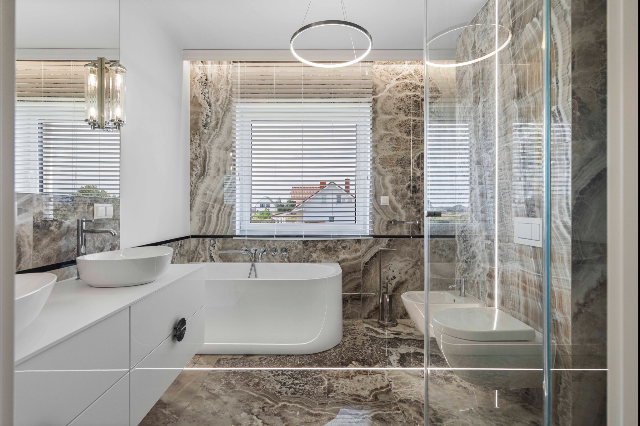 Elegancka łazienka z marmurowymi ścianami, białą wanną i umywalkami. Okno z żaluzjami, nowoczesne oświetlenie. Widok na dom przez okno.
