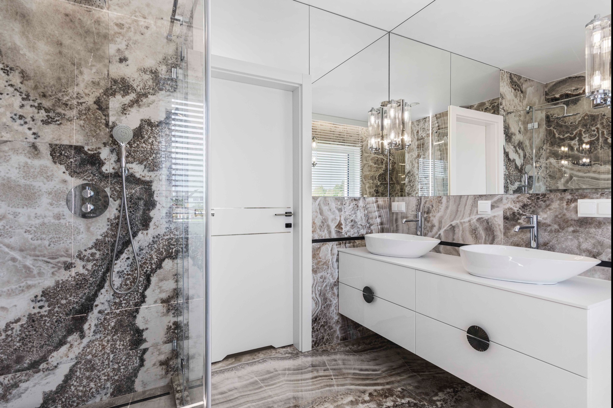 Elegancka łazienka z kabiną prysznicową, umywalkami nablatowymi na białej szafce i lustrem na ścianie z marmurowym wzorem. Nowoczesne oświetlenie.