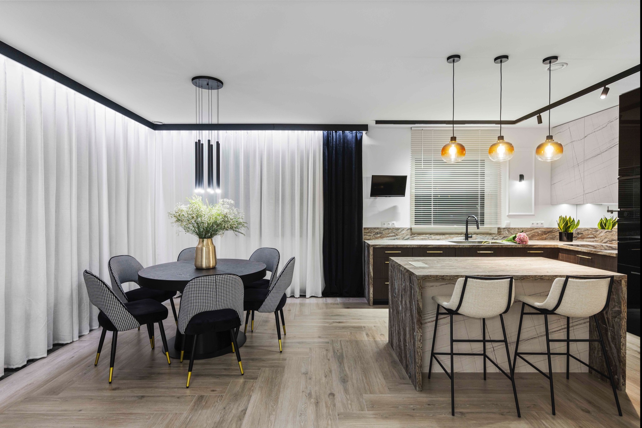Nowoczesne wnętrze: jadalnia z okrągłym stołem, krzesłami w pepitkę i kuchnia z wyspą, oświetlone wiszącymi lampami. Parkiet ułożony w jodełkę.