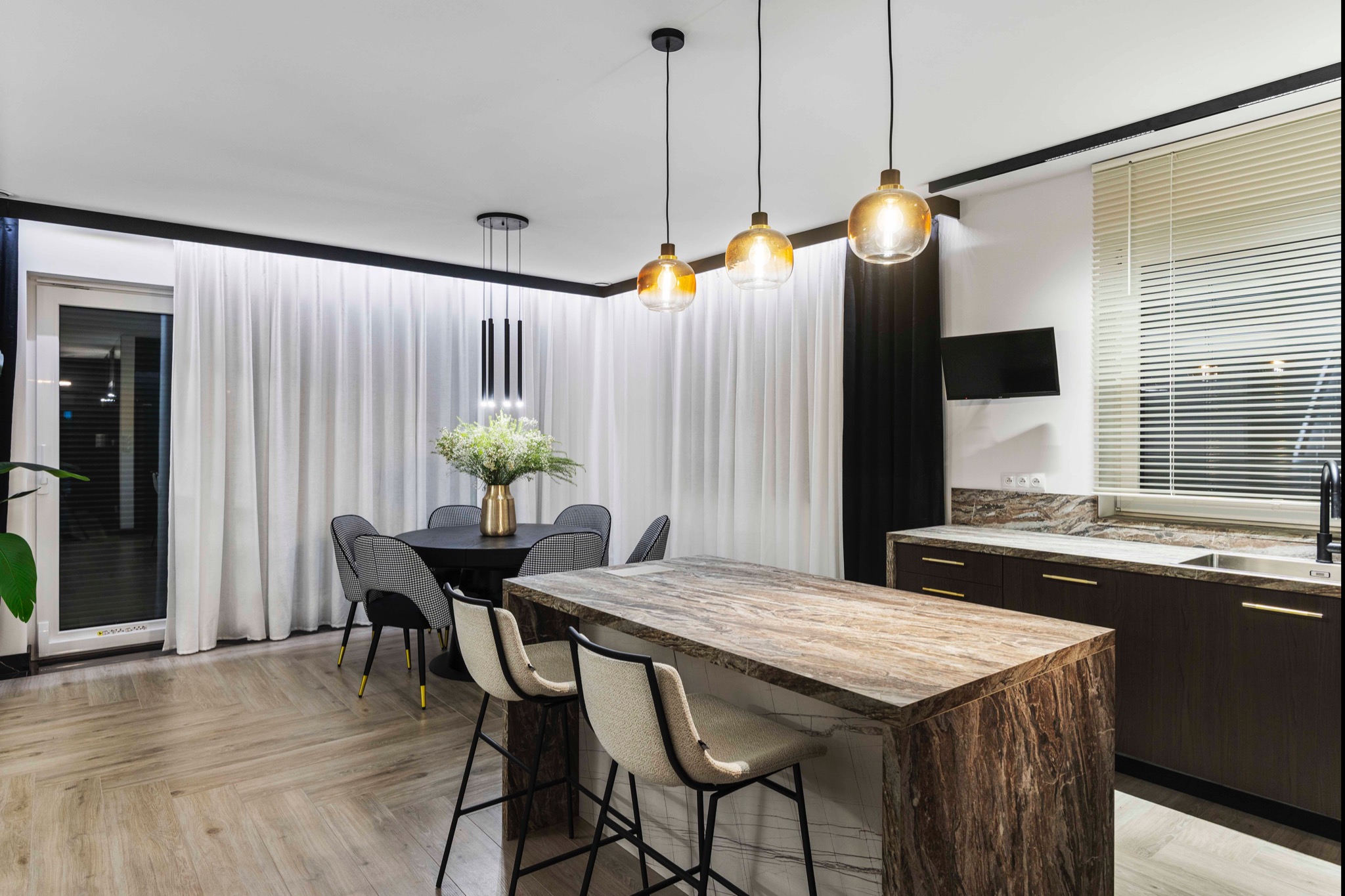 Nowoczesna kuchnia z wyspą o marmurowym blacie, okrągłym stołem i designerskimi lampami wiszącymi. Jasne, przestronne wnętrze z drewnianą podłogą i zasłonami.