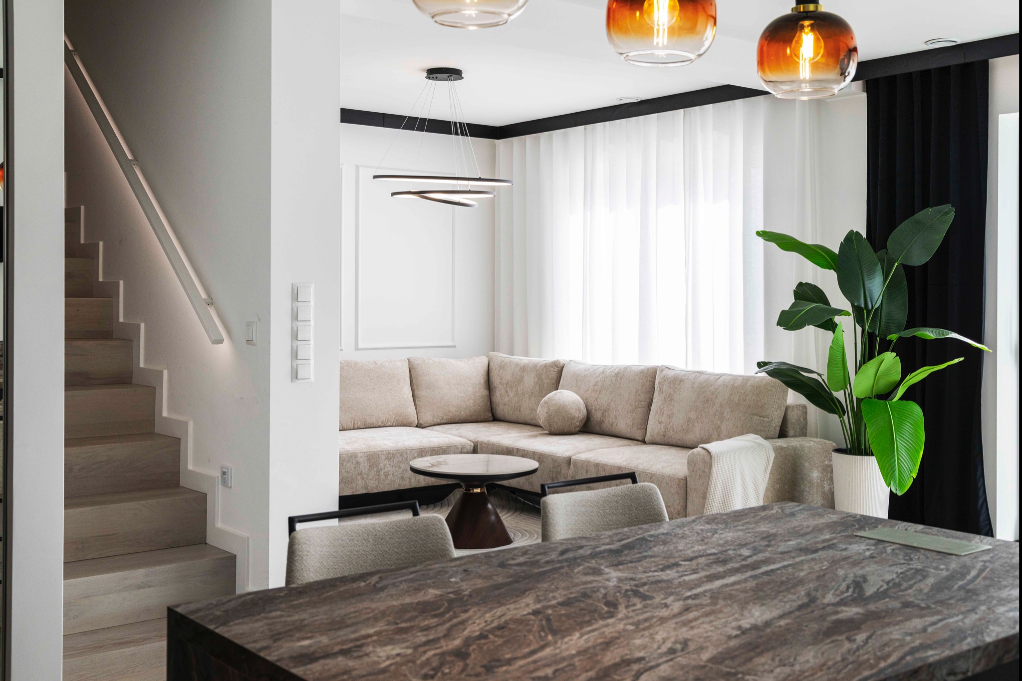 Jasne wnętrze z beżową sofą narożną, drewnianymi schodami z podświetleniem LED i marmurowym blatem stołu na pierwszym planie. Nowoczesne lampy wiszące.