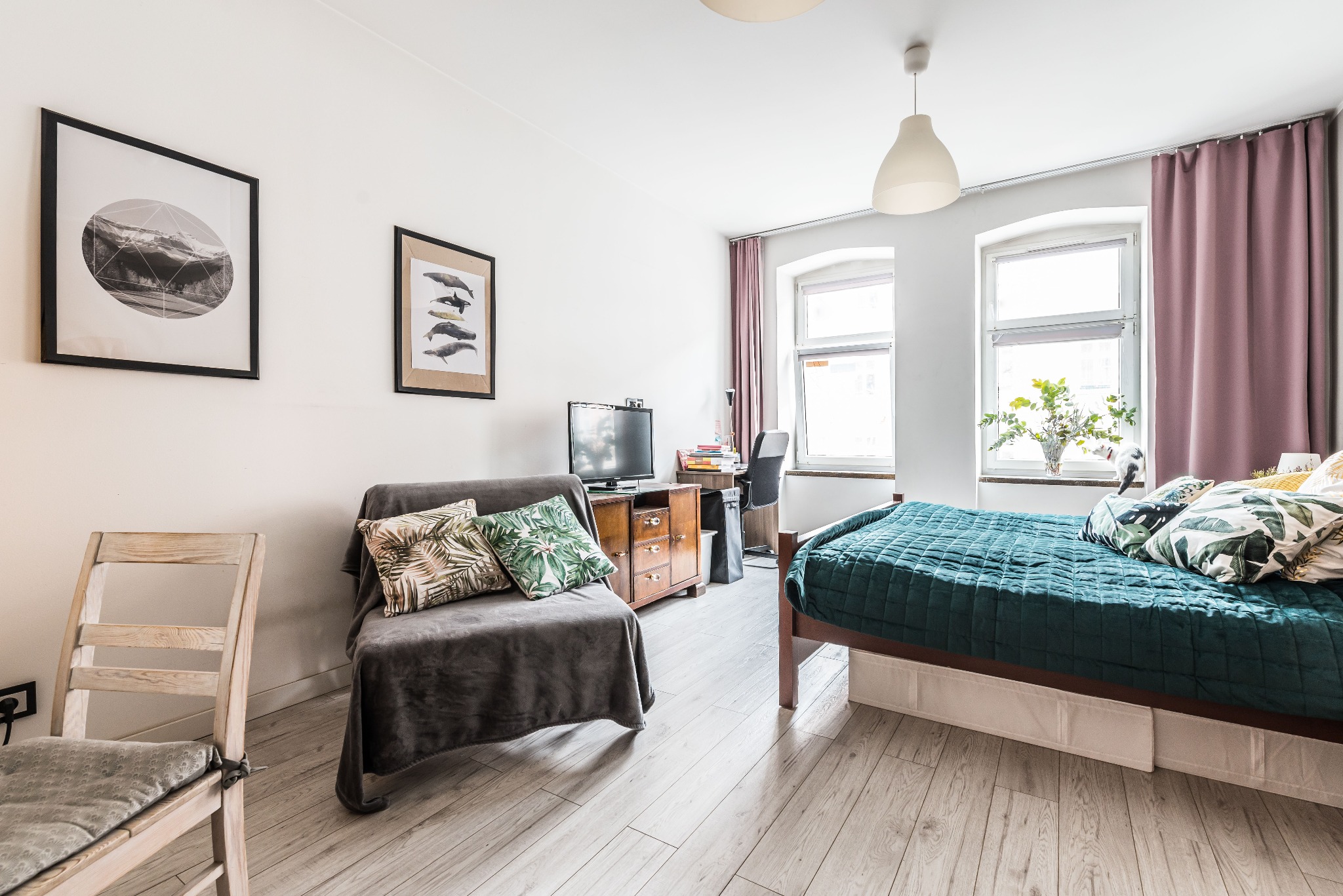 Jasne wnętrze pokoju z drewnianą podłogą, łóżkiem z zieloną narzutą i poduszkami w liście, szarą sofą z poduszkami, biurkiem z komputerem i dwoma oknami z różowymi zasłonami.