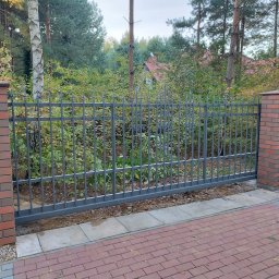 METALO-PLASTYKA JUSTYNA CZYŻ - Przesuwna brama wjazdowa z metalowymi sztachetami i ozdobnymi elementami, zamontowana między murowanymi słupkami z cegły klinkierowej, widok z zewnątrz na tle drzew i krzewów.