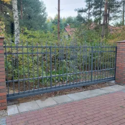 Przesuwna brama wjazdowa z metalowymi sztachetami i ozdobnymi elementami, zamontowana między murowanymi słupkami z cegły klinkierowej, widok z zewnątrz na tle drzew i krzewów.