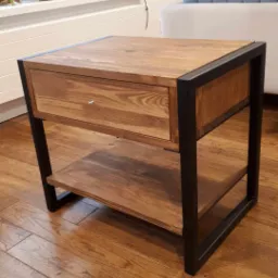 Drewniany stolik nocny z szufladą i metalową ramą na tle drewnianej podłogi, z widocznymi elementami wystroju wnętrza.