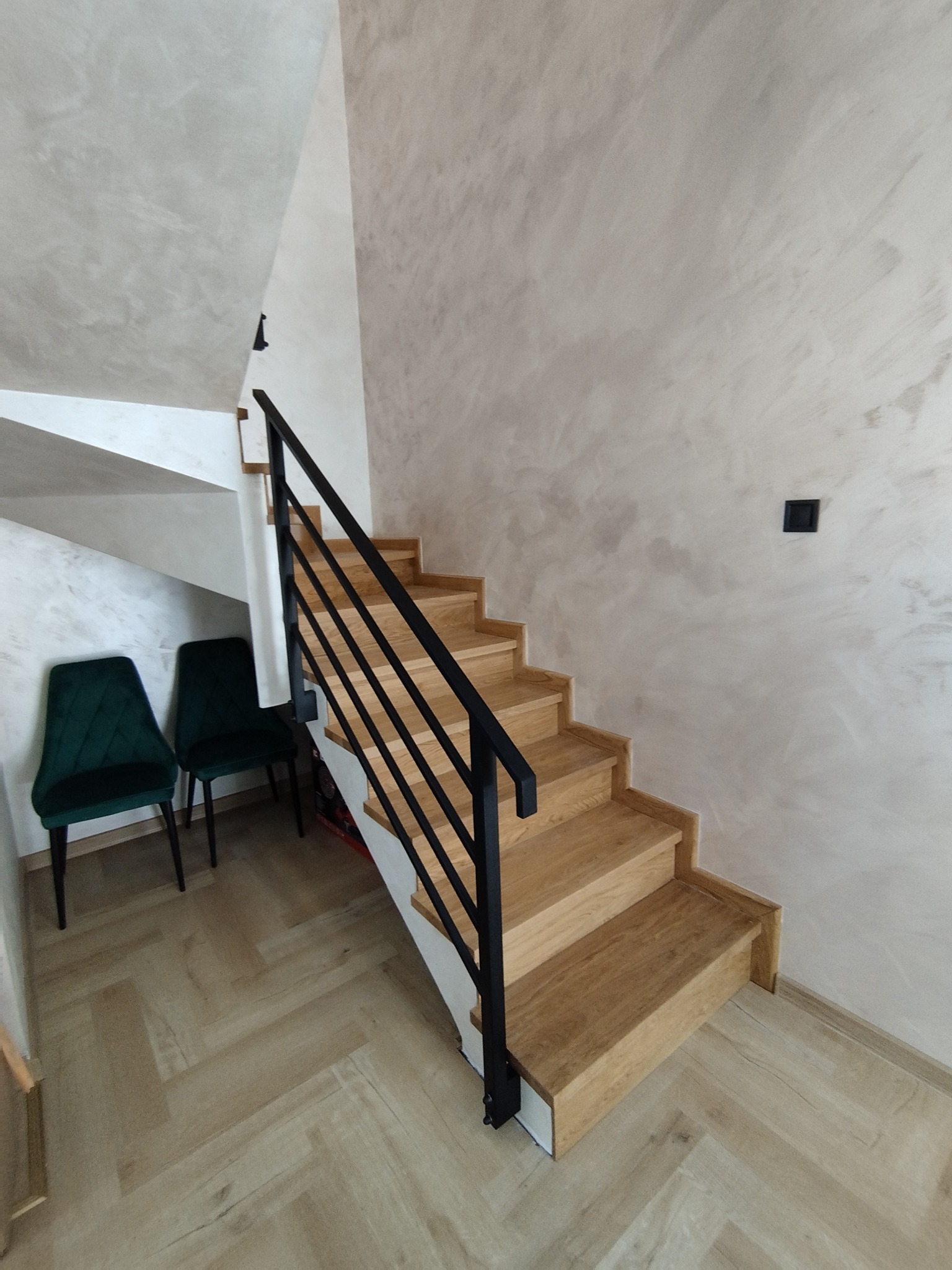 Drewniane schody z czarną, minimalistyczną balustradą, oświetlone naturalnym światłem, z dwoma zielonymi krzesłami w tle i parkietem ułożonym w jodełkę.